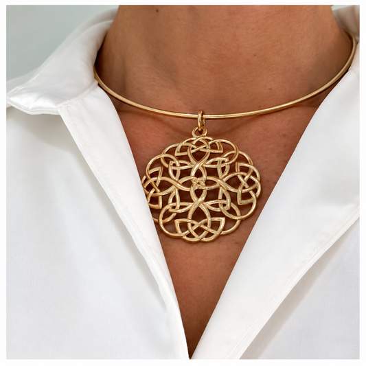 A close-up of a person wearing a delicate gold bangle necklace with a large, intricate Celtic knot pendant, styled with a white collared shirt.