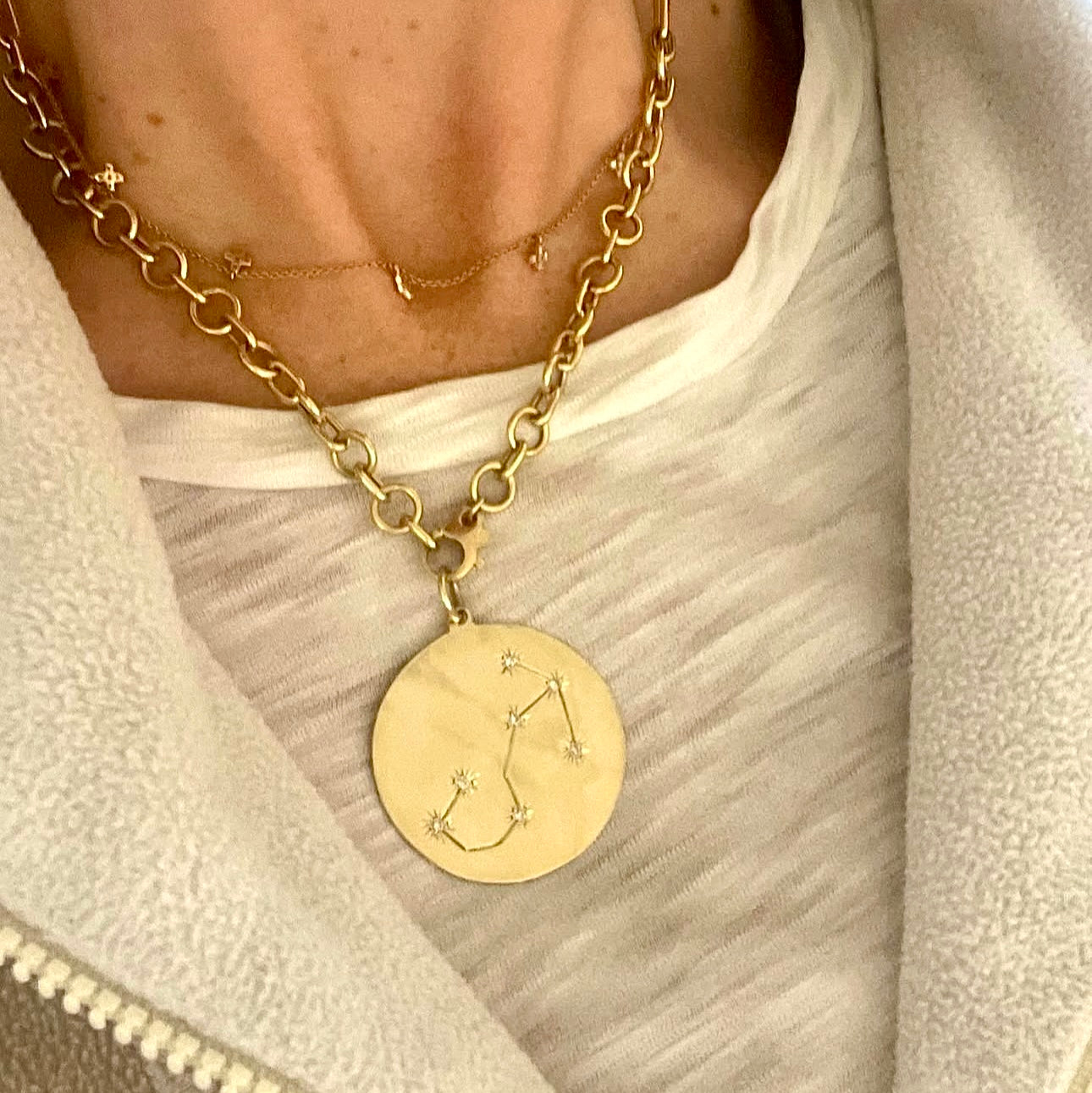 Gold necklace with a circular pendant featuring a constellation design, worn by a person in a light-colored garment.