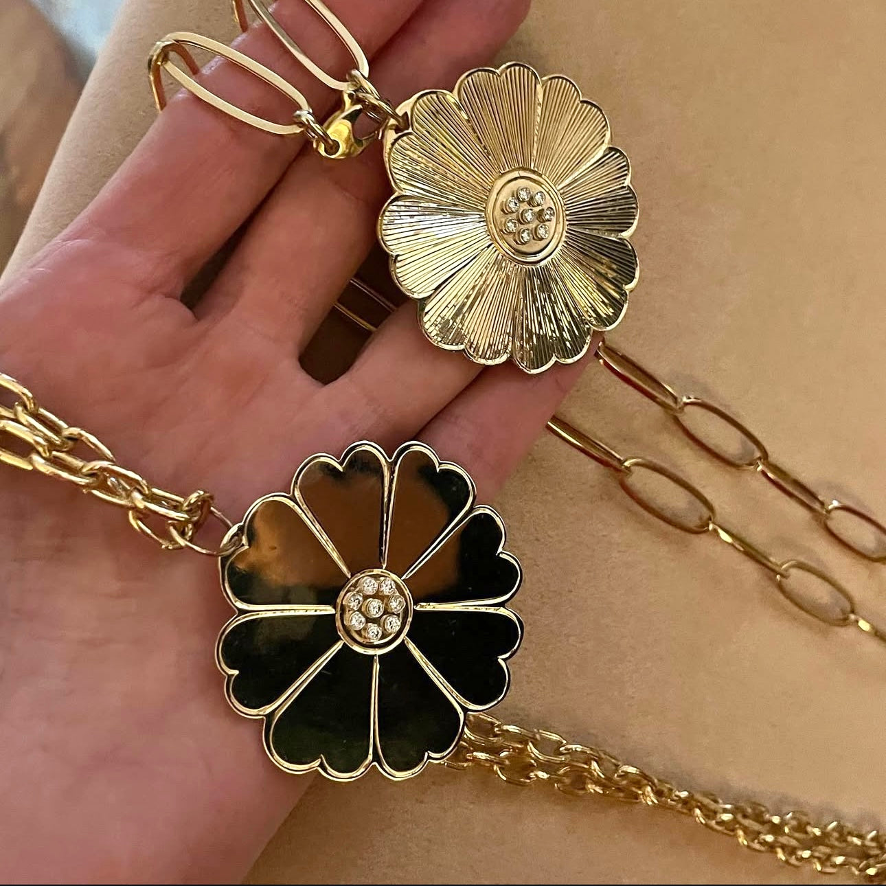 Two gold daisy pendants held in a hand, one is flat, shiny finish, the other is etched.