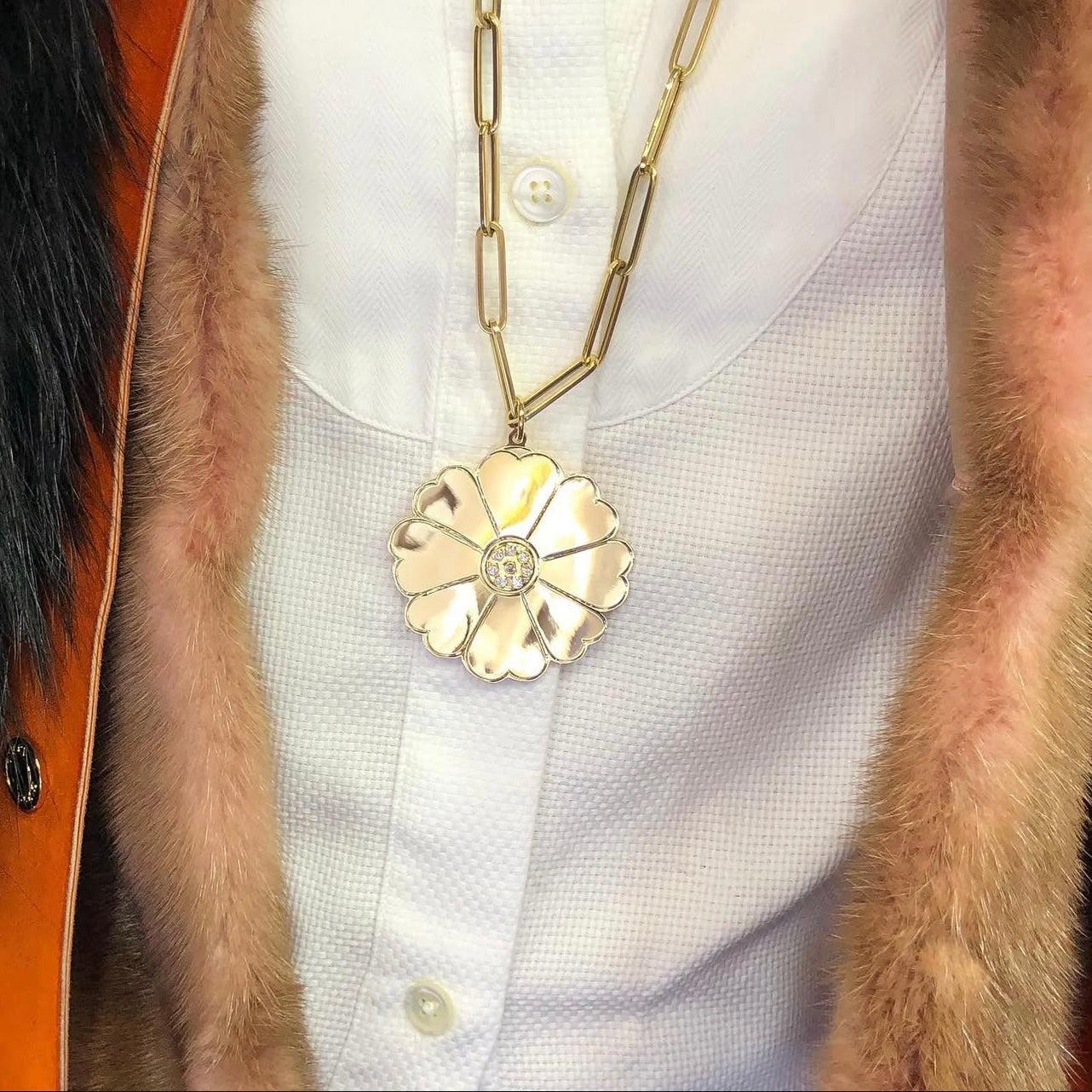Gold necklace with a shiny daisy pendant on a white shirt and fur coat.