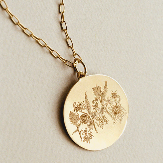 A gold round pendant necklace engraved with delicate wildflowers, resting on a textured light beige surface.