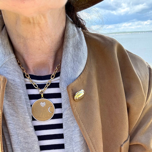 A round gold pendant is worn by a woman on the beach in a cropped photo. The pendant says "I love you to the moon and back" with a heart symbol set with diamonds instead of the word "love" and a crescent moon symbol instead of the word "moon". Gold pendant has a circle of close-set pave diamonds around its diameter, and is shown on a gold "Travel chain", another offered product.