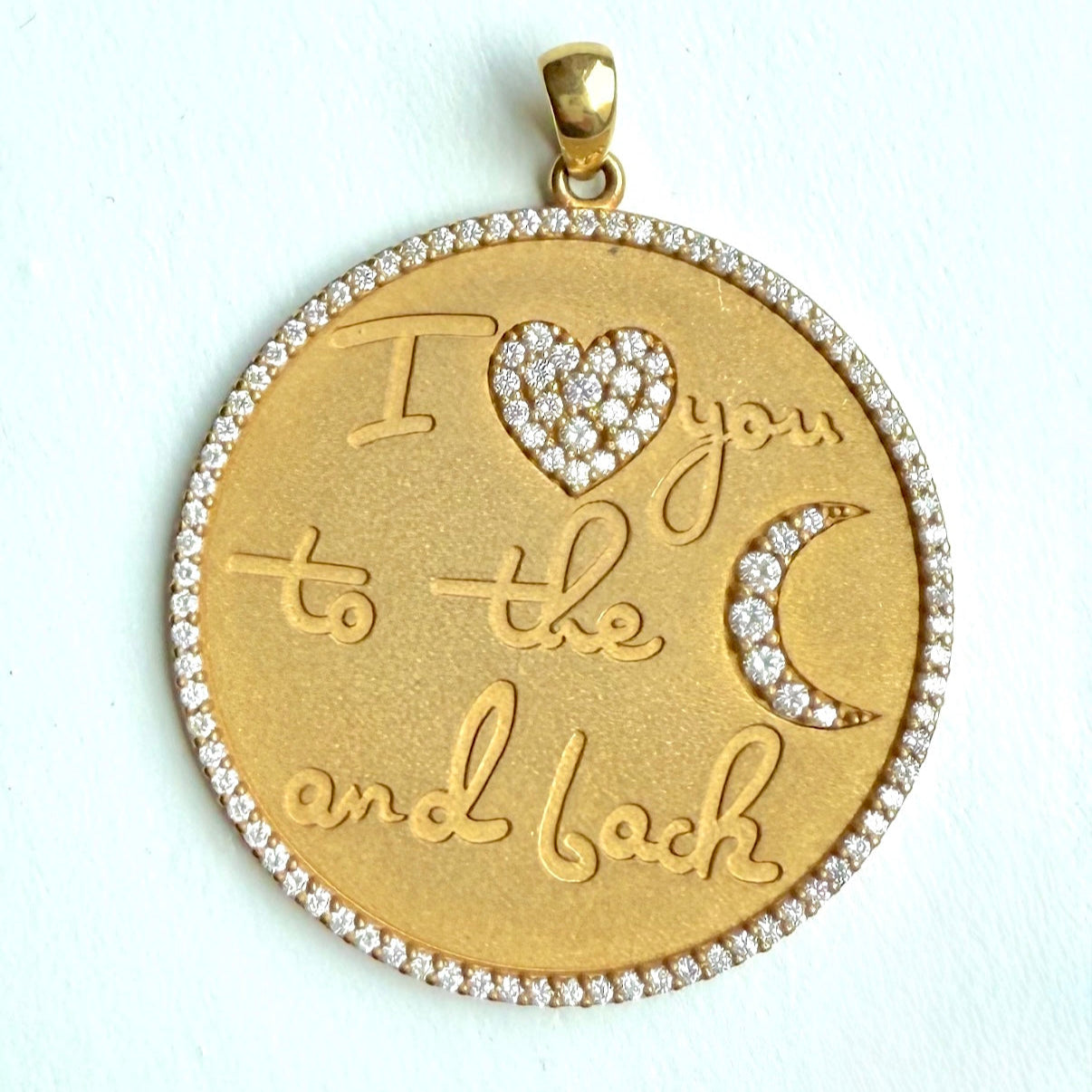 A round gold pendant that says "I love you to the moon and back" with a heart symbol set with diamonds instead of the word "love" and a crescent moon symbol set with diamonds instead of the word "moon". Gold pendant has a circle of close-set pave diamonds around its diameter.