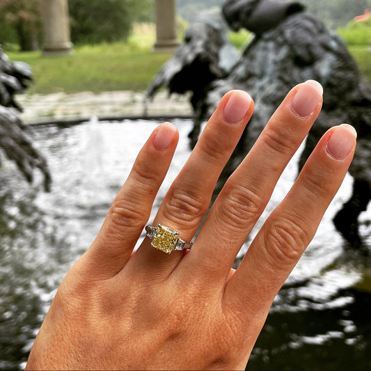 Person's hand with diamond ring in front of blurry outdoor background.