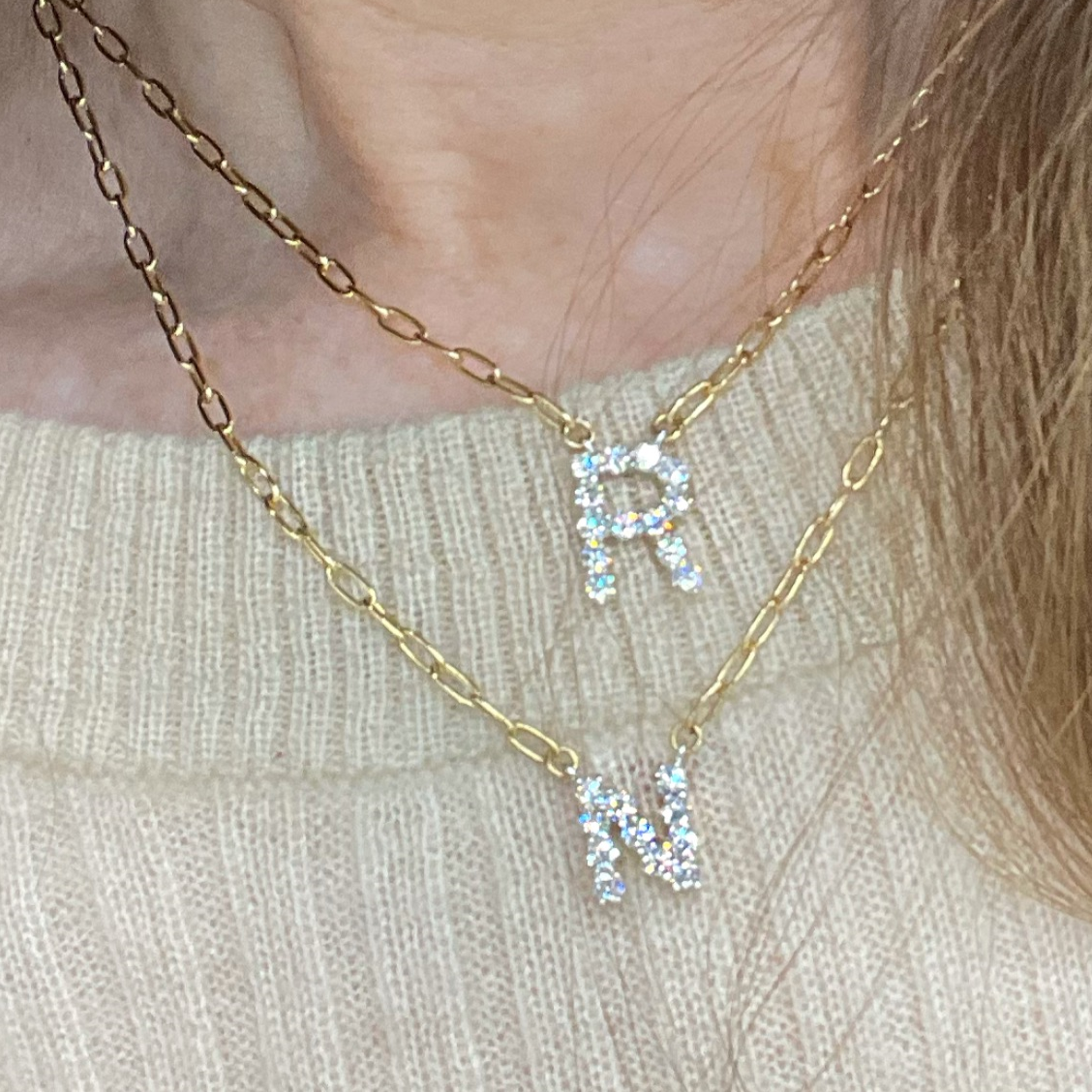 A gold-colored necklace with a customizable initial diamond pendant in the shape of the letter 'R', displayed on a person.