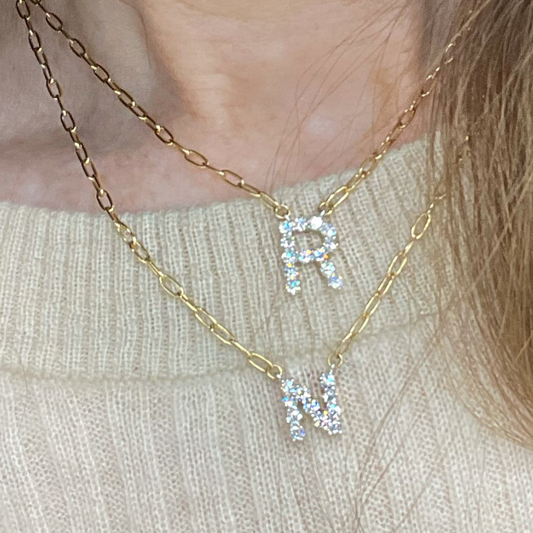 A gold-colored necklace with a customizable initial diamond pendant in the shape of the letter 'R', displayed on a person.