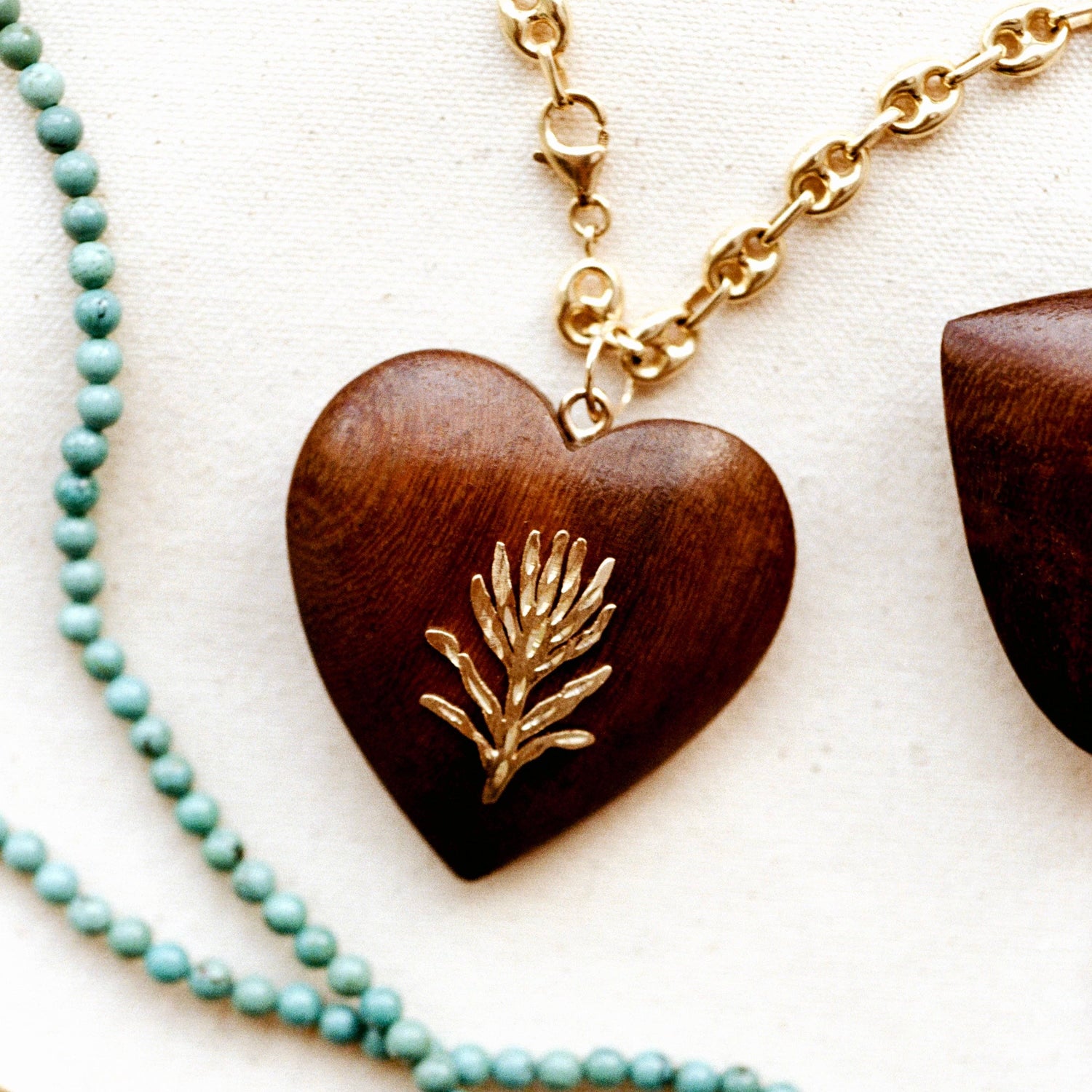 A heart-shaped wooden pendant necklace with a polished gold pine branch inlay, shown on a textured off-white fabric background next to a strand of light blue-green beads.