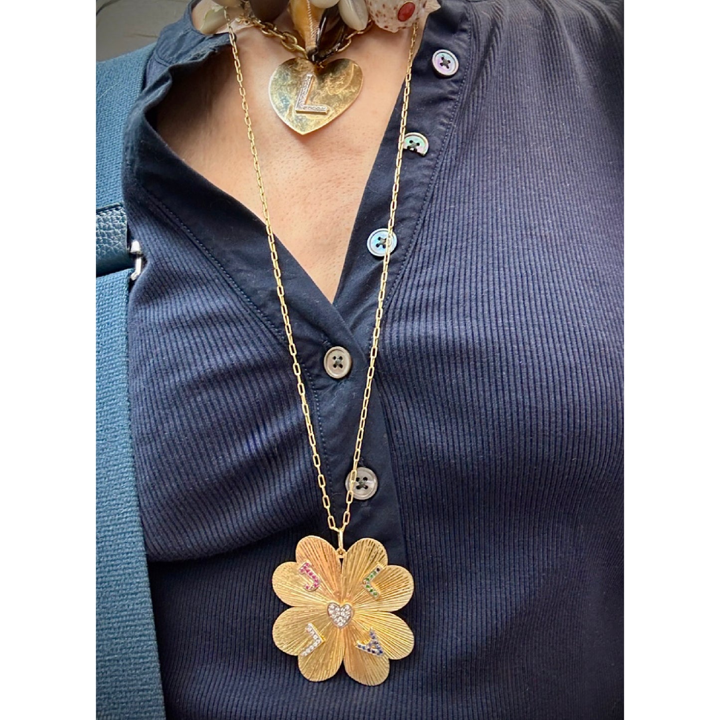 A gold flower-shaped pendant necklace with colorful enamel details and a small heart pendant, worn on a model's neck over a navy blue button-up shirt.