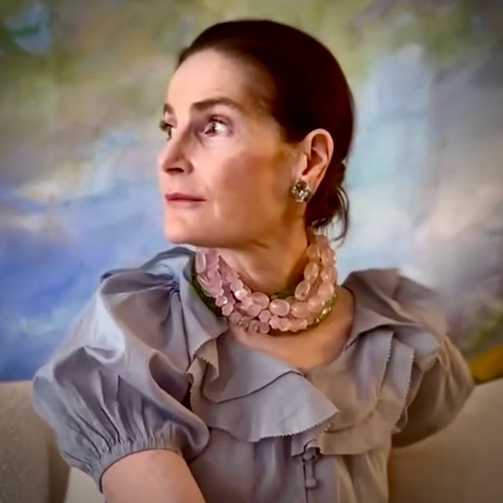 Woman wearing a light purple blouse with ruffled sleeves and a pink necklace against a blurred natural background.