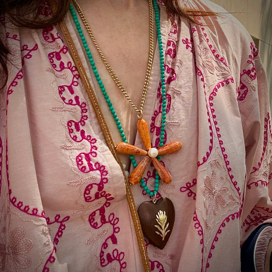 A close-up of a person wearing layered bohemian-style necklaces over a light pink embroidered tunic. The necklaces include a gold chain with a shell flower pendant, a turquoise beaded necklace, and a dark brown heart pendant with gold detailing.