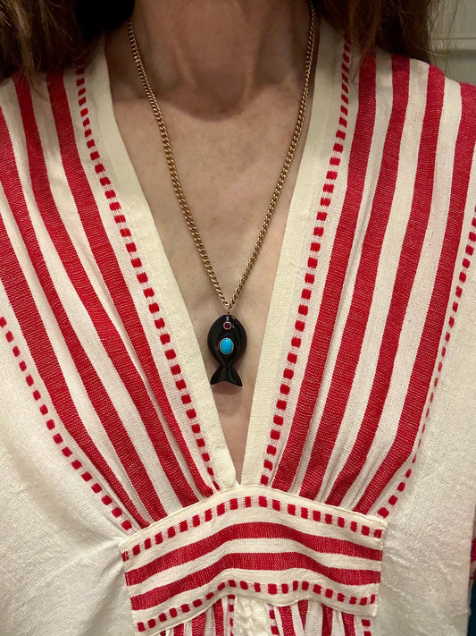 A close-up of a person wearing a gold chain necklace with a black fish-shaped pendant featuring a turquoise blue center, styled over a red and white striped knit top.
