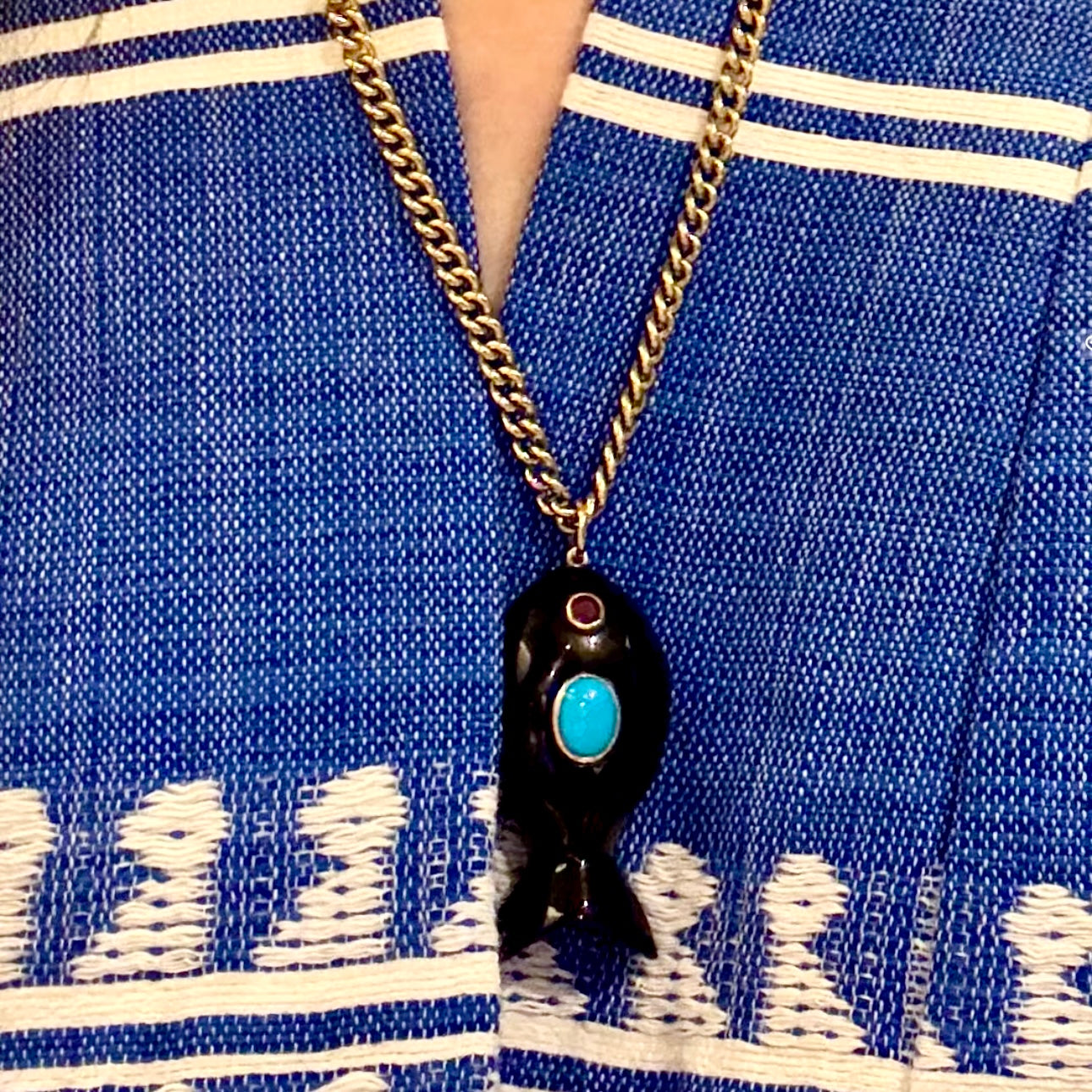 A close-up of a person wearing a black fish-shaped pendant necklace with a turquoise center and gold chain, styled over a blue and white patterned knit top.