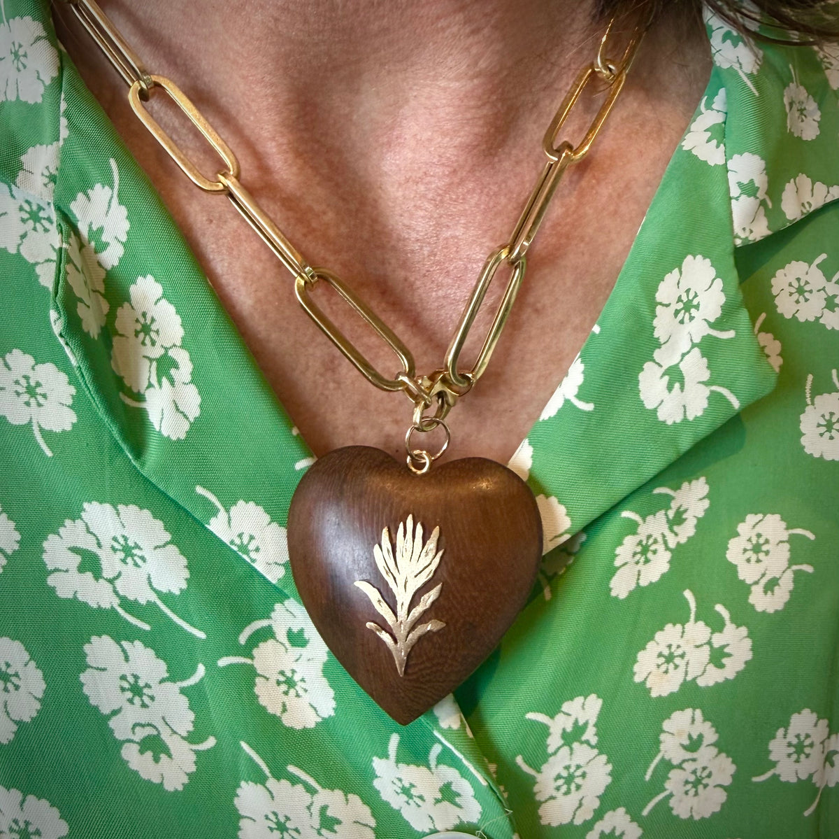 A close-up of a person wearing a gold paperclip chain necklace with a dark brown wooden heart pendant featuring a white floral engraving, paired with a green and white floral shirt.
