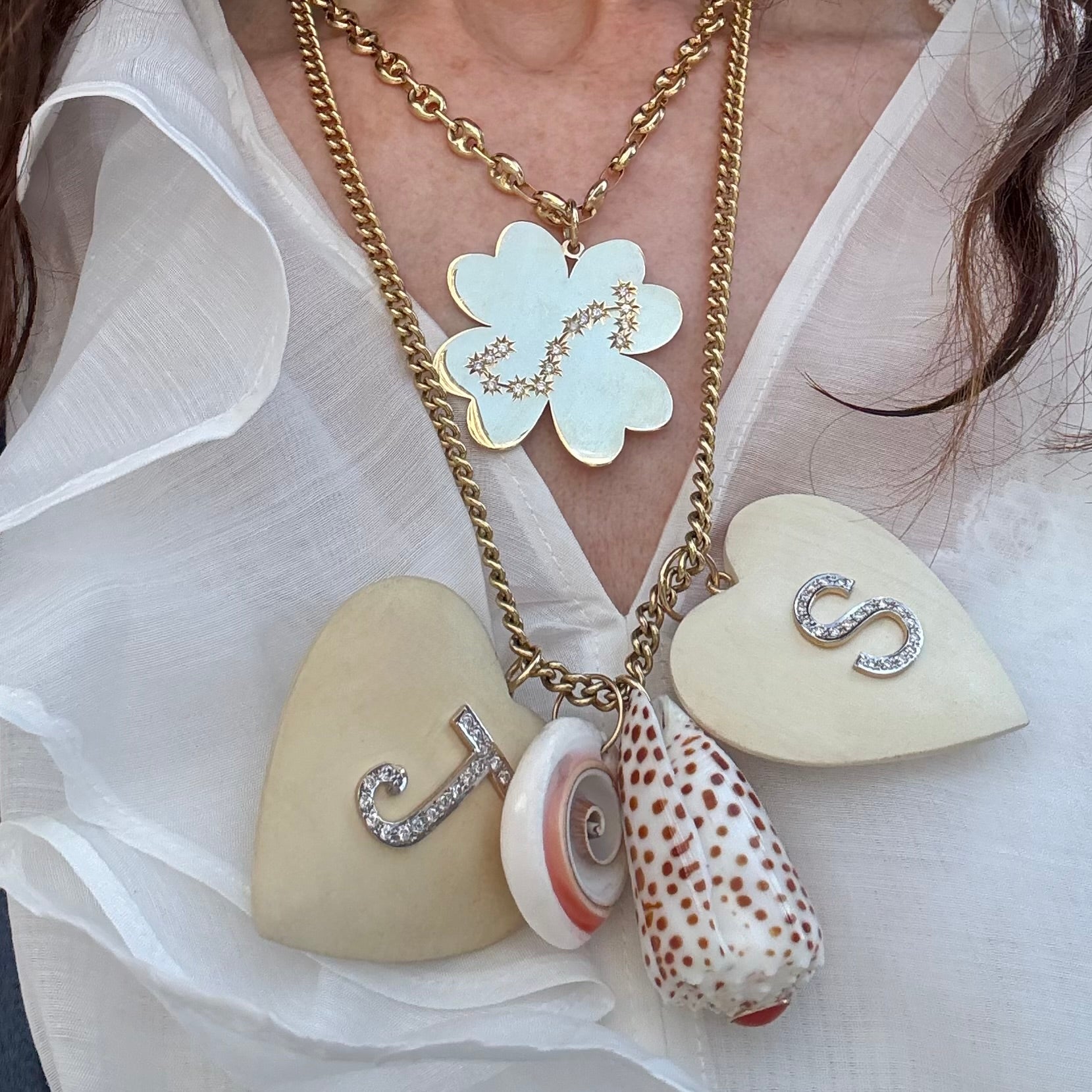 A close-up of a layered necklace worn on a model's neck. The necklace features a gold chain with a large gold clover pendant, a gold chain with two cream-colored heart pendants (one with a diamond 'S' and one with a 'J'), a small white and pink seashell, and a larger patterned seashell with brown spots.