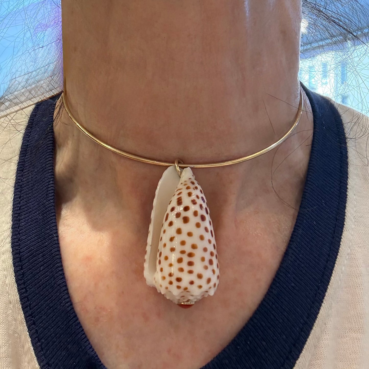 A close-up of a person's neck wearing a thin gold choker necklace with a large white cone shell pendant decorated with brown spots.