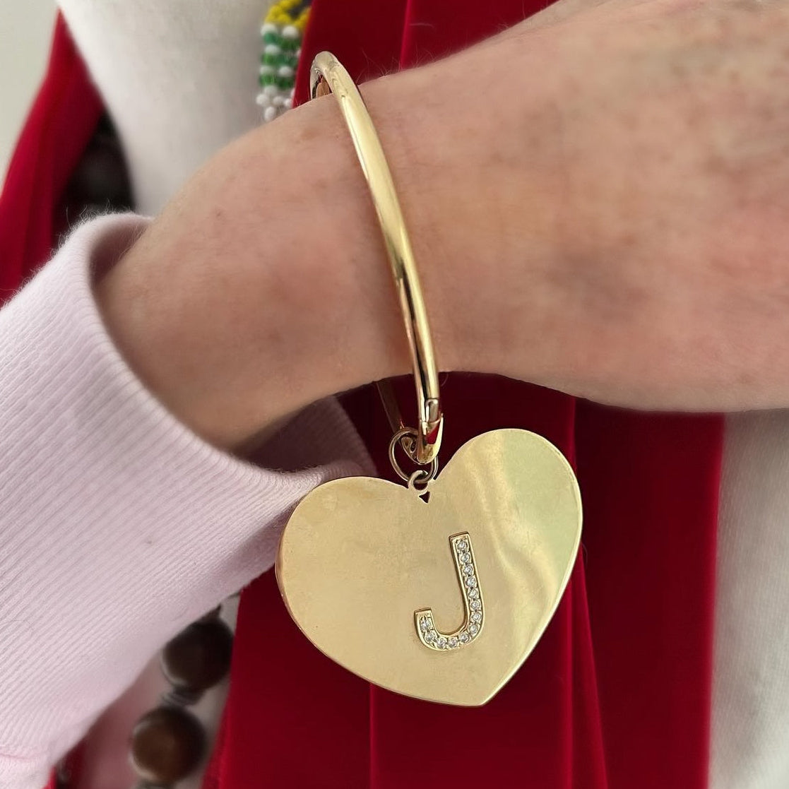 Gold heart-shaped bracelet with a letter 'J' charm on a wrist against a red fabric background.