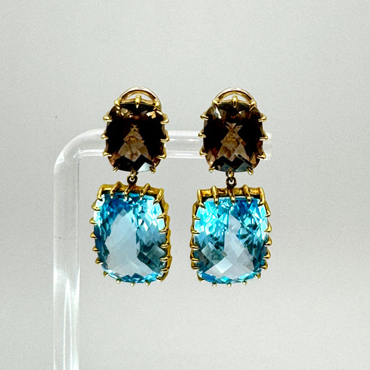 Pair of earrings with smoky topaz and blue topaz stones on a clear stand against a light gray background.