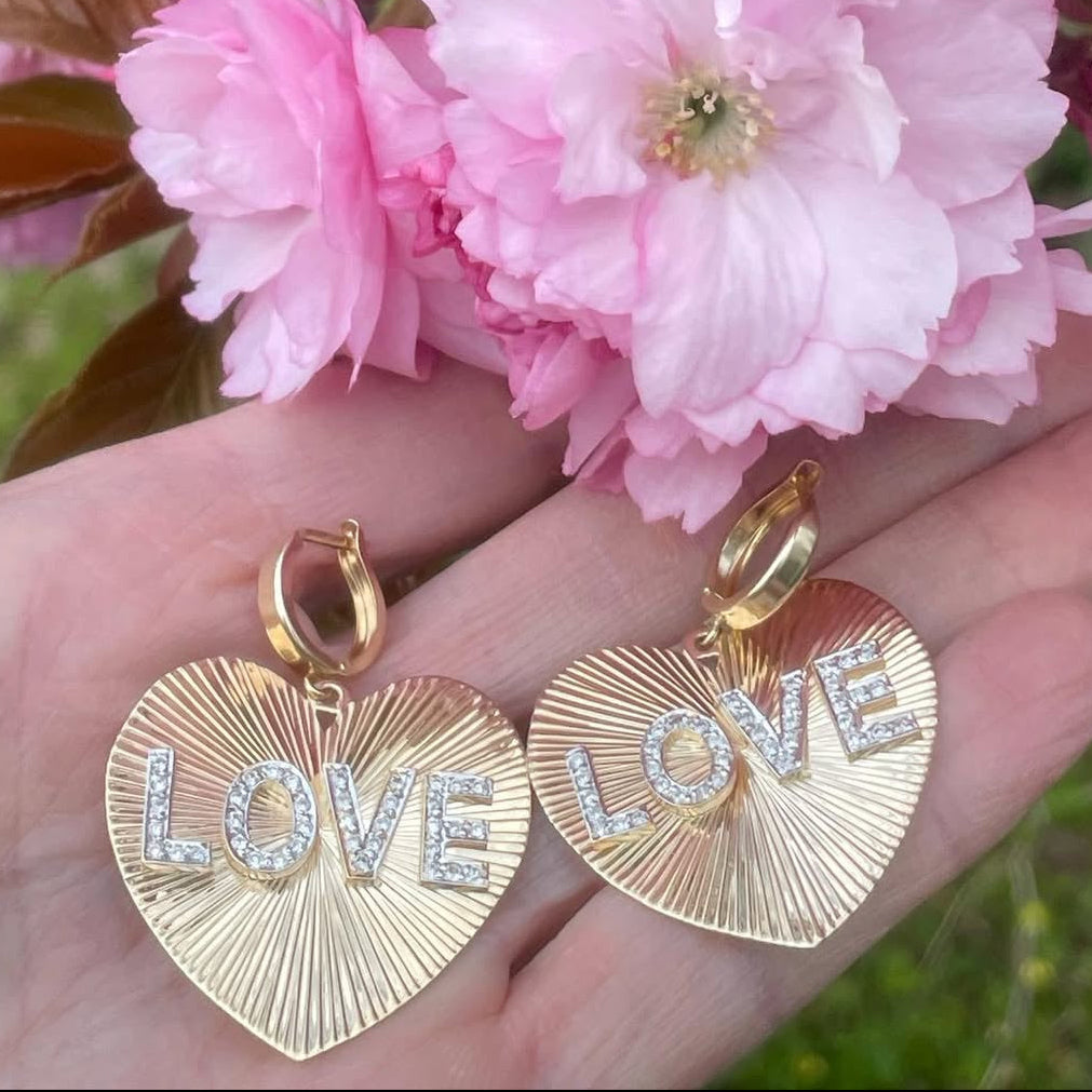 Gold 'LOVE' heart-shaped earrings held against pink cherry blossoms.