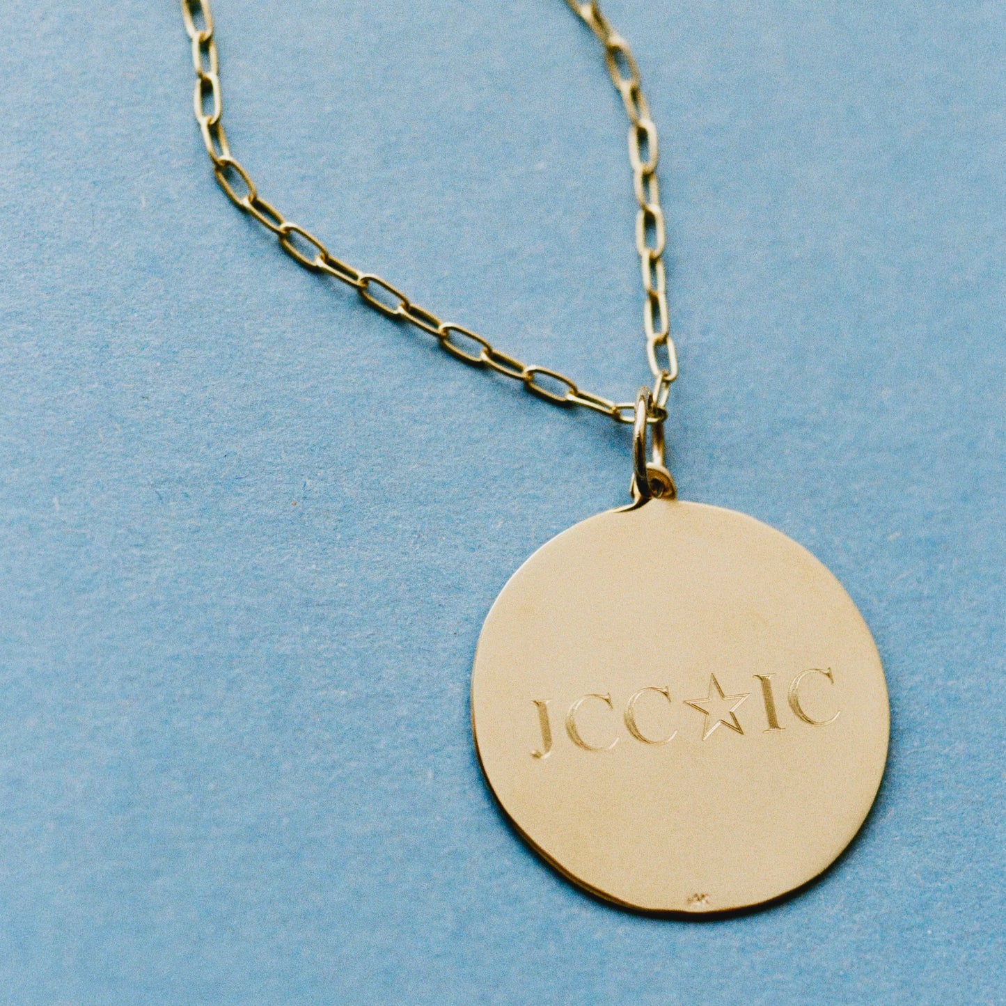 A gold round disc pendant necklace engraved with the initials 'JCC-IC' and a star, hanging from a gold paperclip chain against a textured light blue background.
