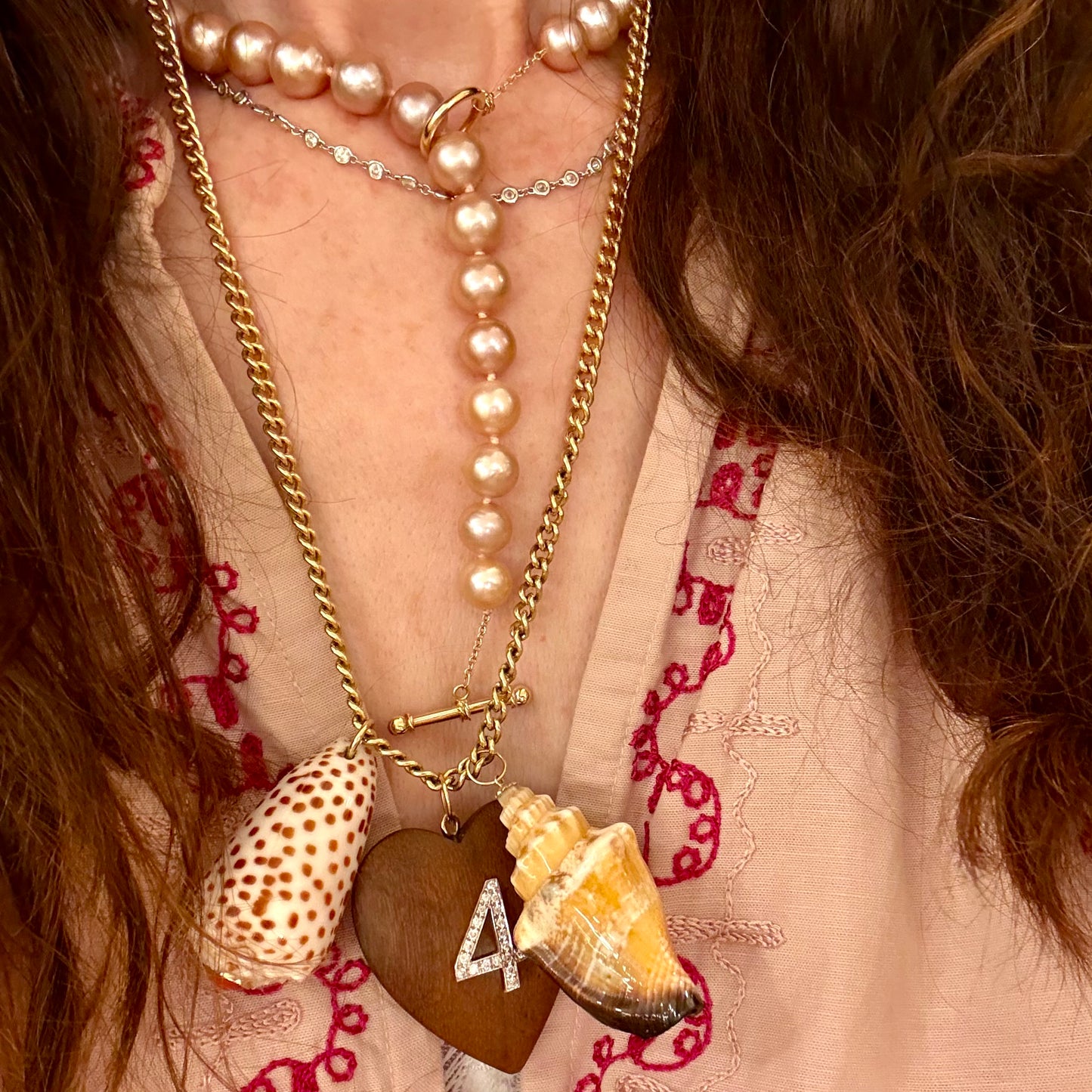 A close-up of a person wearing layered necklaces, including gold-tone chains with conch shell and wooden heart pendants, pearl beads, and a rhinestone-embellished necklace.