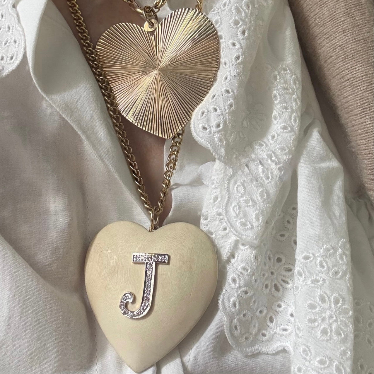 A close-up of a person wearing layered necklaces over a white embroidered blouse. One necklace has a large cream heart pendant with a silver letter 'J' and a gold chain, while another necklace above it has a gold heart pendant with sunburst detailing.