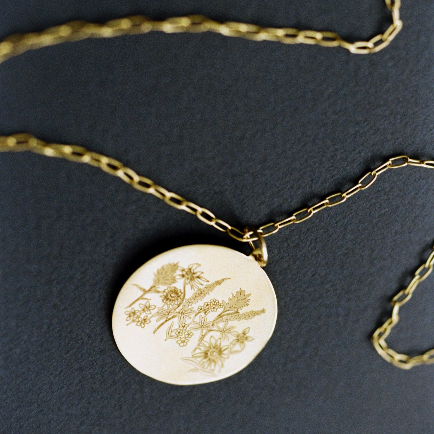 A gold chain necklace with a round gold disc pendant featuring a delicate botanical engraving, displayed on a dark textured background.