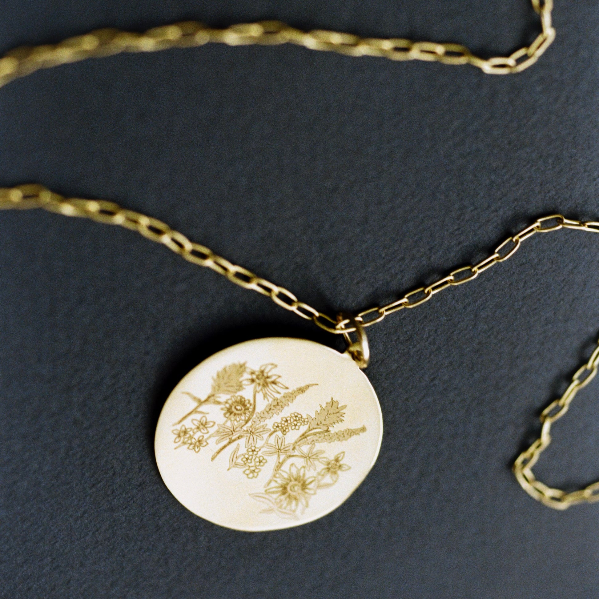A gold chain necklace with a round gold disc pendant featuring a delicate botanical engraving, displayed on a dark textured background.