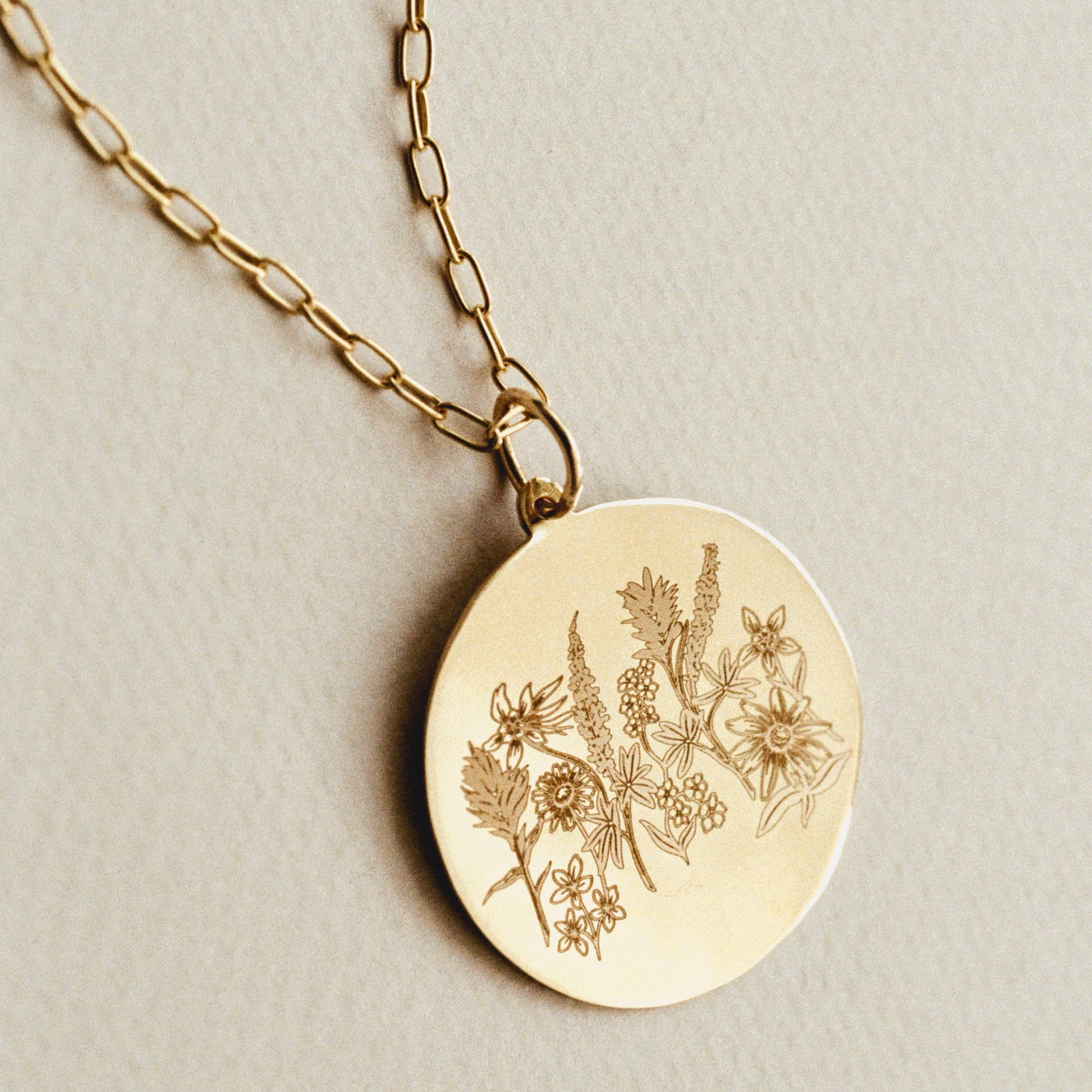 A gold round pendant necklace engraved with delicate wildflowers, resting on a textured light beige surface.