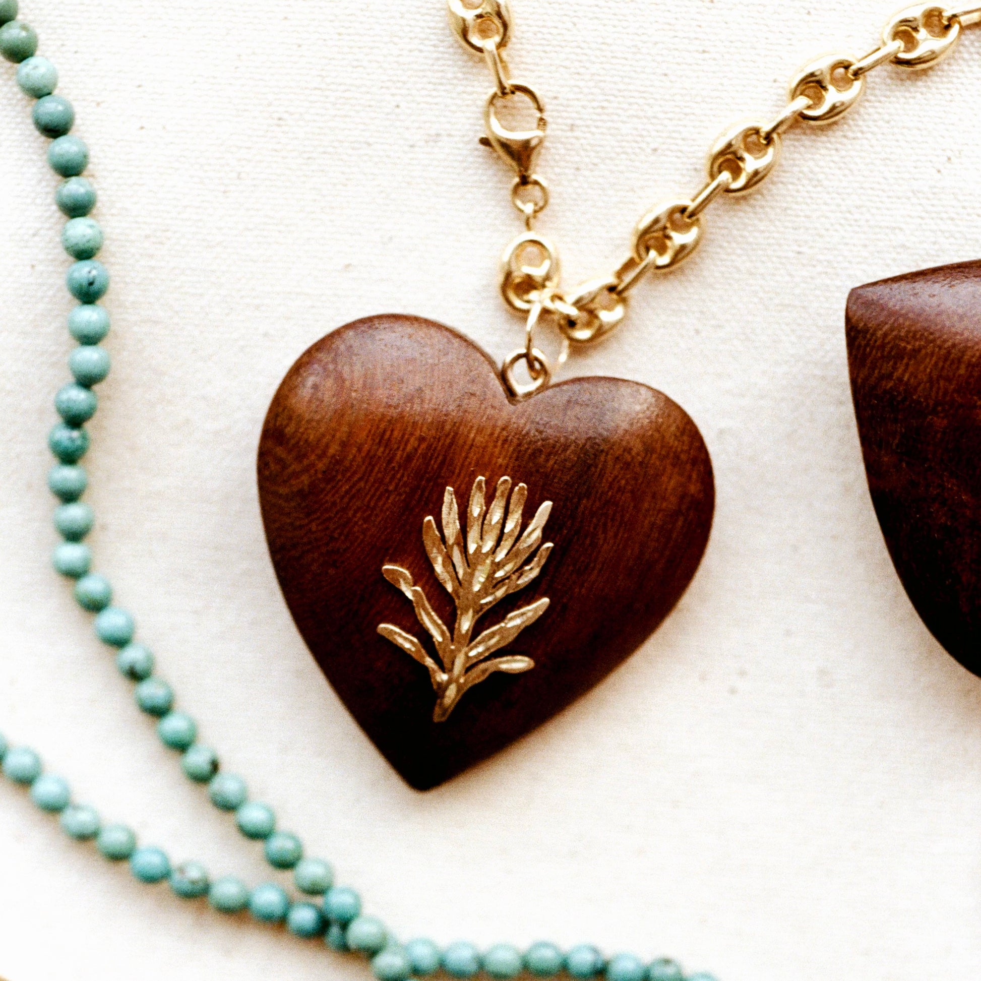 A heart-shaped wooden pendant necklace with a polished gold pine branch inlay, shown on a textured off-white fabric background next to a strand of light blue-green beads.
