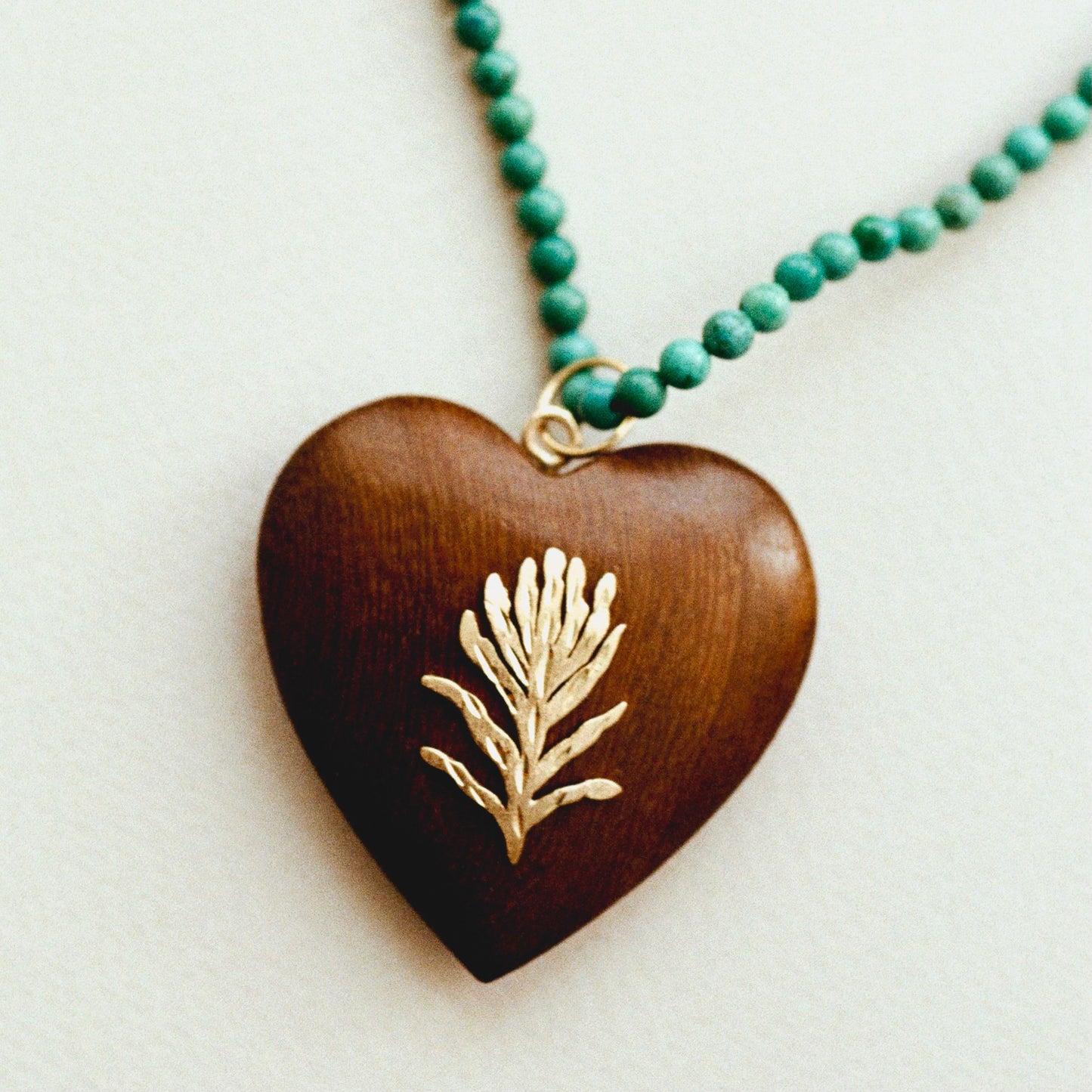 A heart-shaped wooden pendant necklace with a gold-toned floral design in the center, hanging from a green beaded chain on a light background.
