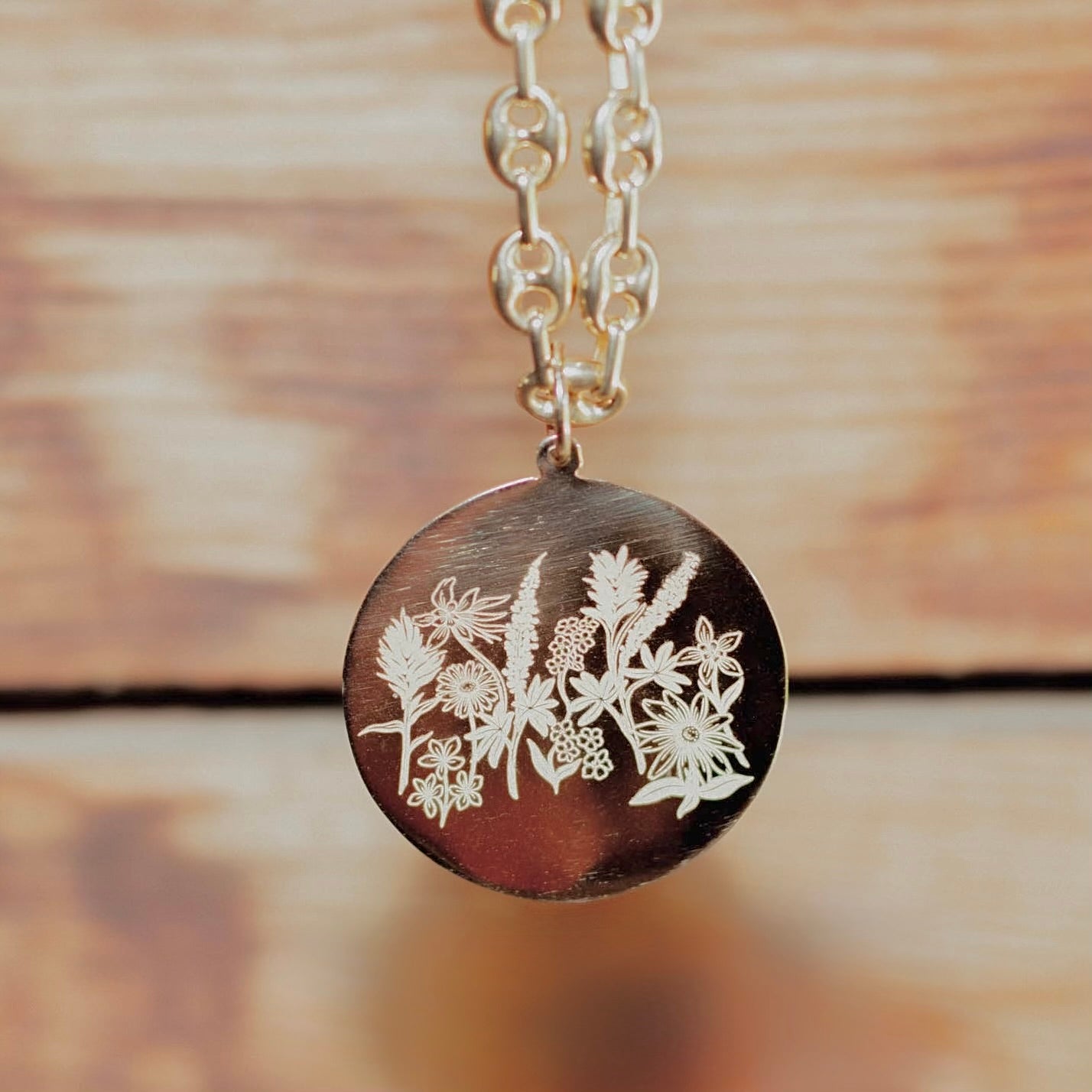 A close-up of a round gold pendant necklace featuring engraved wildflower patterns, hanging from a gold-toned chain against a blurred wooden background.