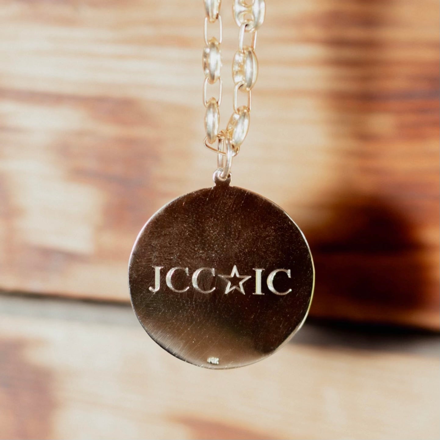 A close-up of a round gold pendant necklace engraved with the letters 'JCC' and a star followed by 'IC', hanging from a chain against a blurred wooden background.