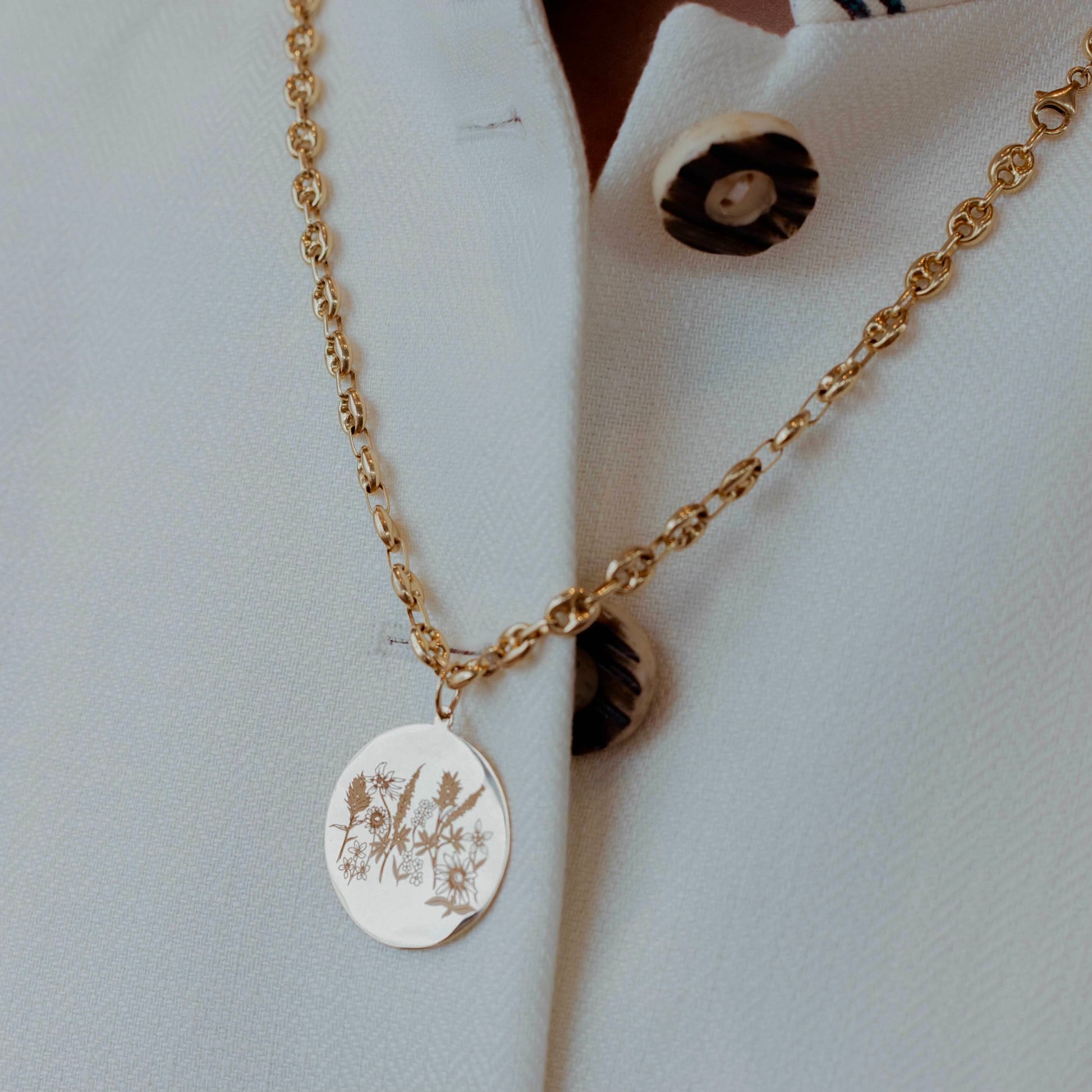 A gold chain necklace with a round gold pendant engraved with floral details, worn on a white shirt with brown buttons.