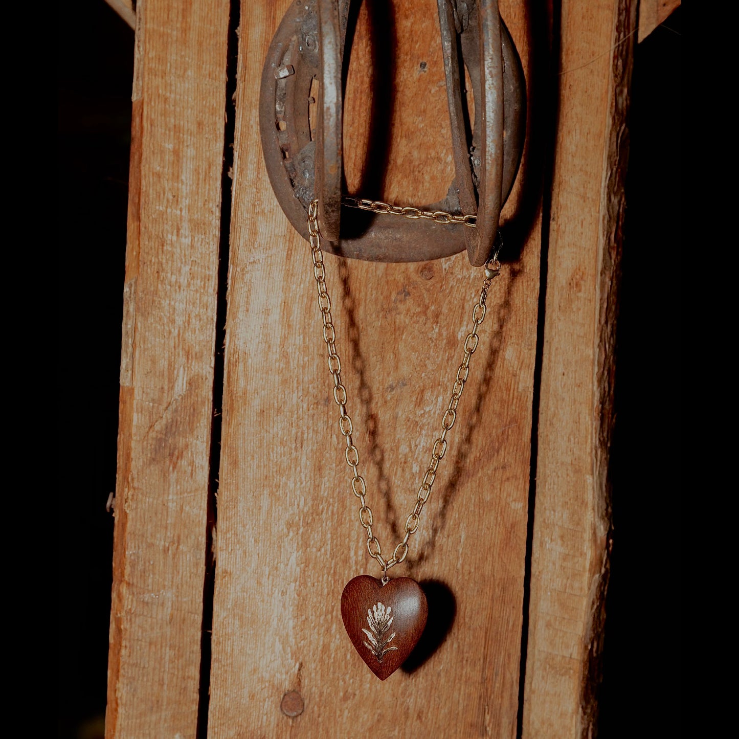 A rustic brown wooden heart pendant necklace with a silver leaf engraving, hanging from a gold-toned chain and a weathered horseshoe against a vertical wooden plank background.