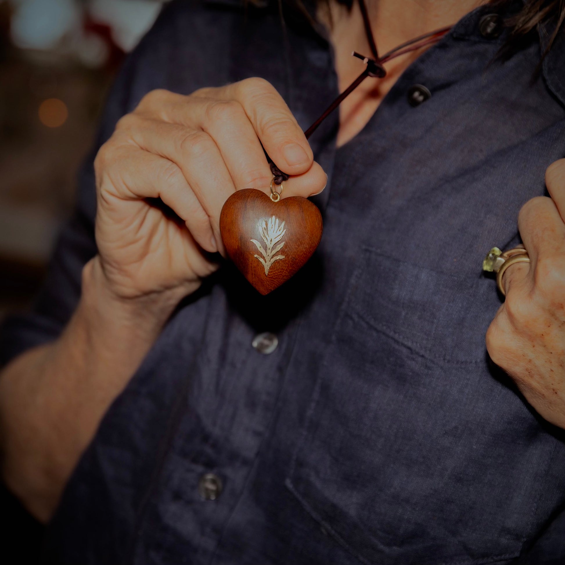 A person wearing a dark navy button-up shirt holds a small carved wooden heart pendant with a white leaf design and a gold-toned loop, shown against a softly blurred background.