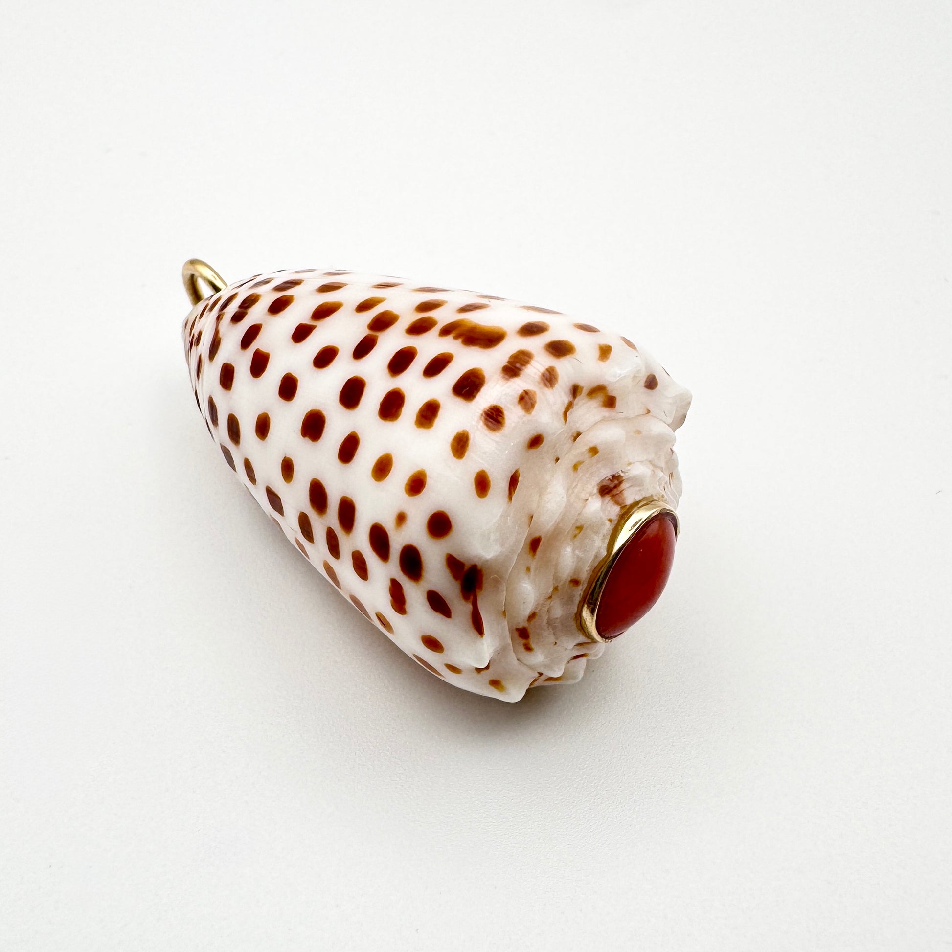 A cone-shaped shell pendant with brown spots on a white base and a coral cabochon stone set in a gold bezel, photographed on a plain white background.