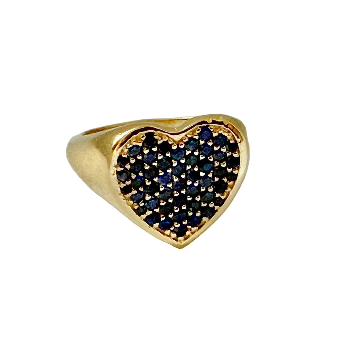 A gold signet-style ring with a heart-shaped face paved with small dark blue stones, photographed on a light neutral background.